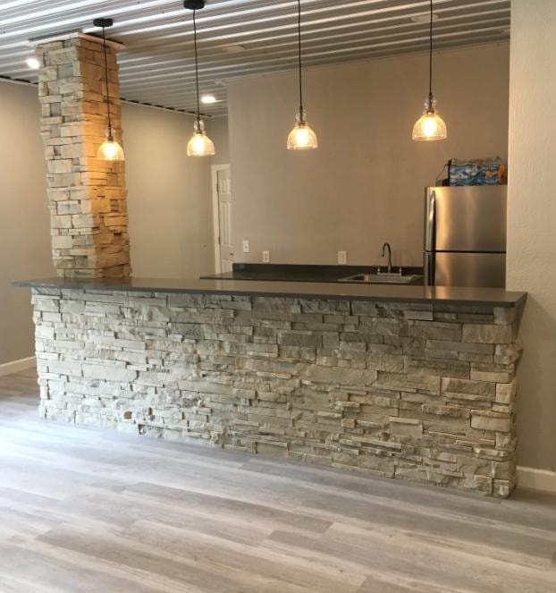 Basement bar build with stacked stone surround, stacked stone column, pendant lighting, and stainless refrigerator